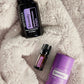 Three doTERRA Serenity products arranged on a soft white faux-fur surface: a large black bottle, a small amber dropper bottle, and a purple stick container. doTERRA Serenity Sleep System