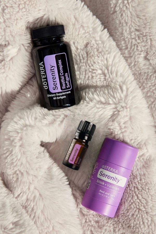 Three doTERRA Serenity products arranged on a soft white faux-fur surface: a large black bottle, a small amber dropper bottle, and a purple stick container. doTERRA Serenity Sleep System
