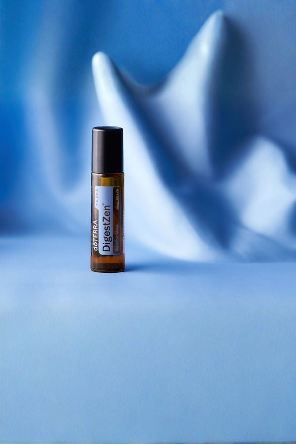 A small amber glass roller bottle with a black cap and a white label reading 'doTERRA DigestZen Touch DigestZen' stands on a smooth blue surface against a softly draped blue background.