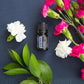 A small amber essential oil bottle labeled 'Pleasant Valley' is arranged on a dark blue textured surface with white and pink flowers and green leaves around it. doTERRA Juniper Berry Essential Oil