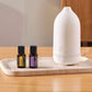 A white essential oil diffuser sits on a light stone tray next to two small amber bottles on a wooden table. dōTERRA LaLuz Diffuser
