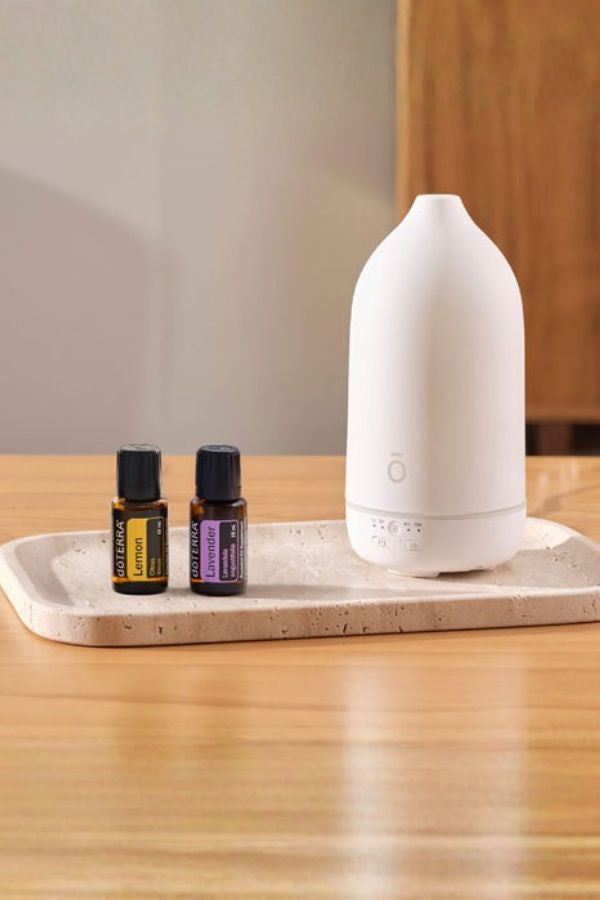 A white essential oil diffuser sits on a light stone tray next to two small amber bottles on a wooden table. dōTERRA LaLuz Diffuser
