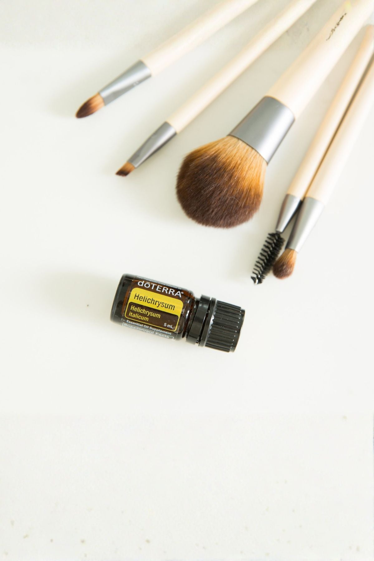 A set of makeup brushes with light wood handles and silver ferrules arranged on a white surface, alongside a small black doTERRA essential oil bottle labeled Decadentia. doTERRA Helichrysum Essential Oil