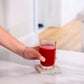 A hand holding a clear glass filled with bright red juice, resting on a square concrete coaster on a white marble countertop in a bright kitchen setting. MetaPWR Recharge Electrolytes - Wild Strawberry