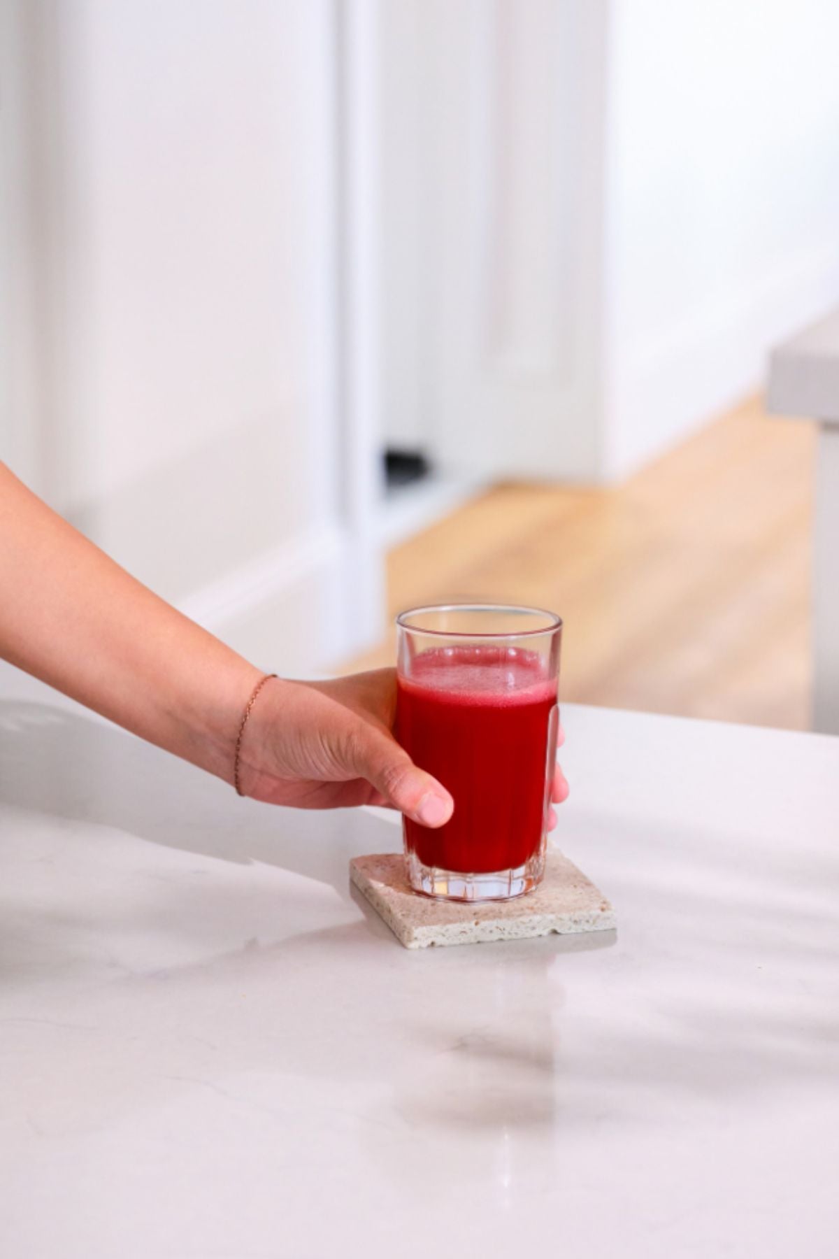 A hand holding a clear glass filled with bright red juice, resting on a square concrete coaster on a white marble countertop in a bright kitchen setting. MetaPWR Recharge Electrolytes - Wild Strawberry