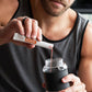 A man in a gray tank top mixes a red liquid into a clear shaker bottle with a black sleeve, holding a white and orange MicroPlenty packet above the bottle. MetaPWR Recharge Electrolytes - Wild Strawberry