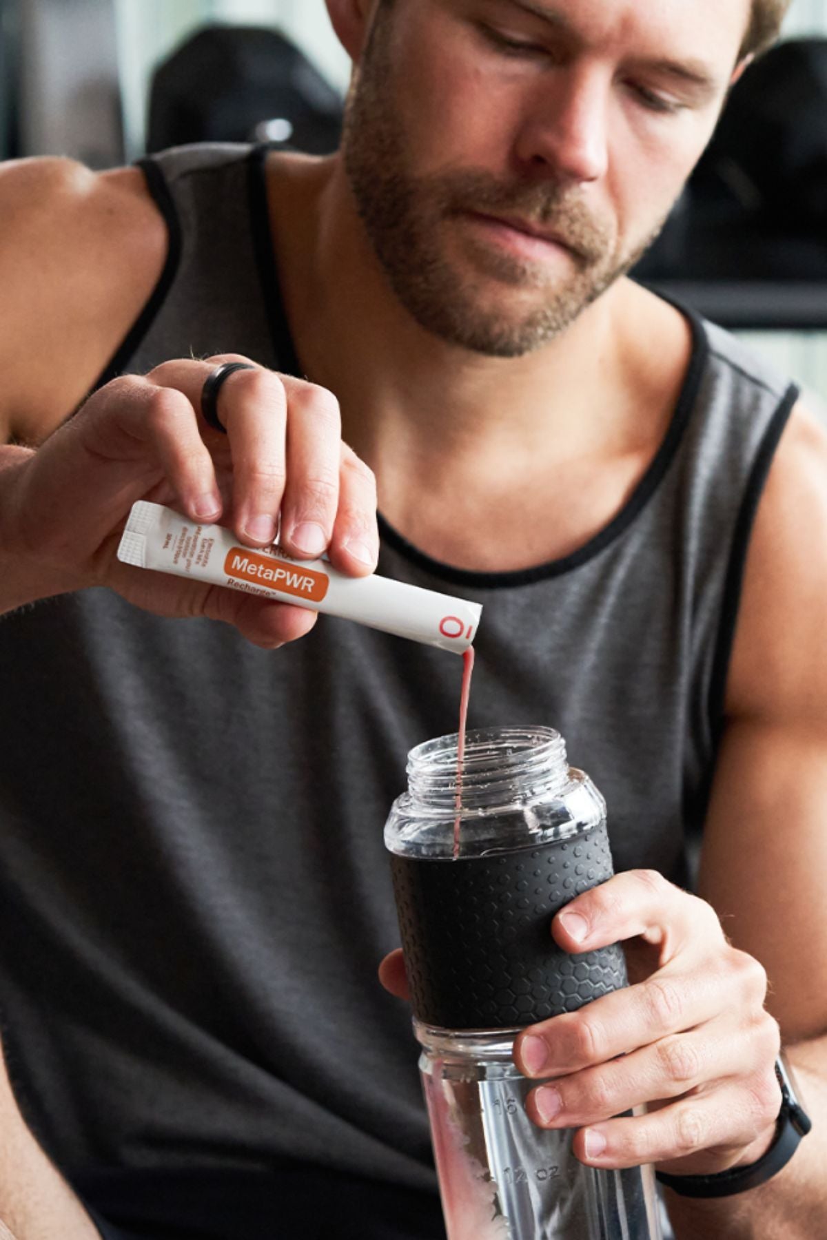 A man in a gray tank top mixes a red liquid into a clear shaker bottle with a black sleeve, holding a white and orange MicroPlenty packet above the bottle. MetaPWR Recharge Electrolytes - Wild Strawberry