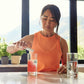A woman in an orange sleeveless top stands at a kitchen counter pouring a pink drink into a glass, with two clear glasses and a bright window view of mountains in the background. MetaPWR Recharge Electrolytes - Wild Strawberry