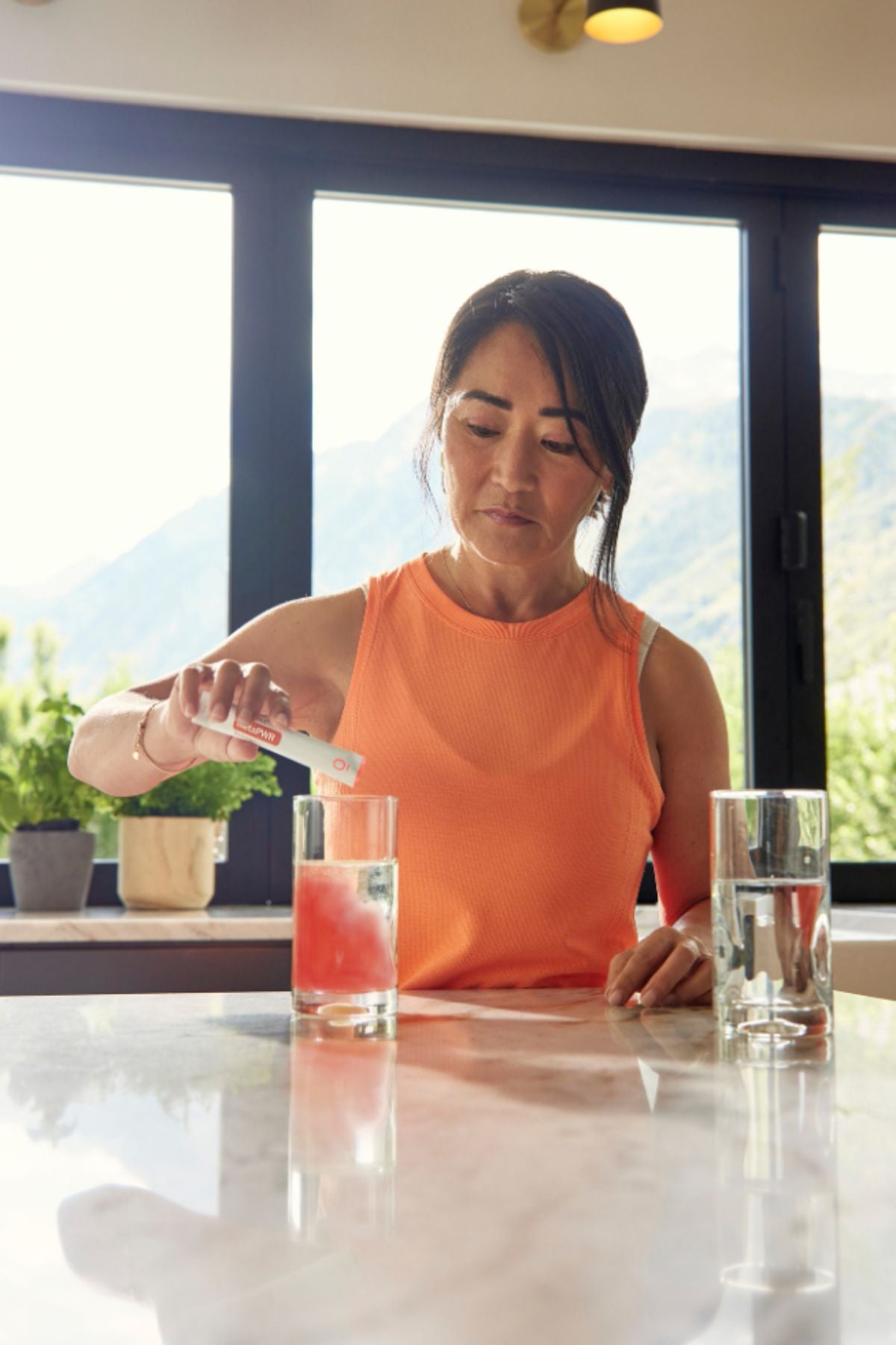 A woman in an orange sleeveless top stands at a kitchen counter pouring a pink drink into a glass, with two clear glasses and a bright window view of mountains in the background. MetaPWR Recharge Electrolytes - Wild Strawberry