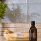 A bottle of doTERRA Castor Oil Wrap Pack oil stands on a countertop next to a folded doTERRA pouch, with a wooden brush in a bowl in the background against a tiled wall.