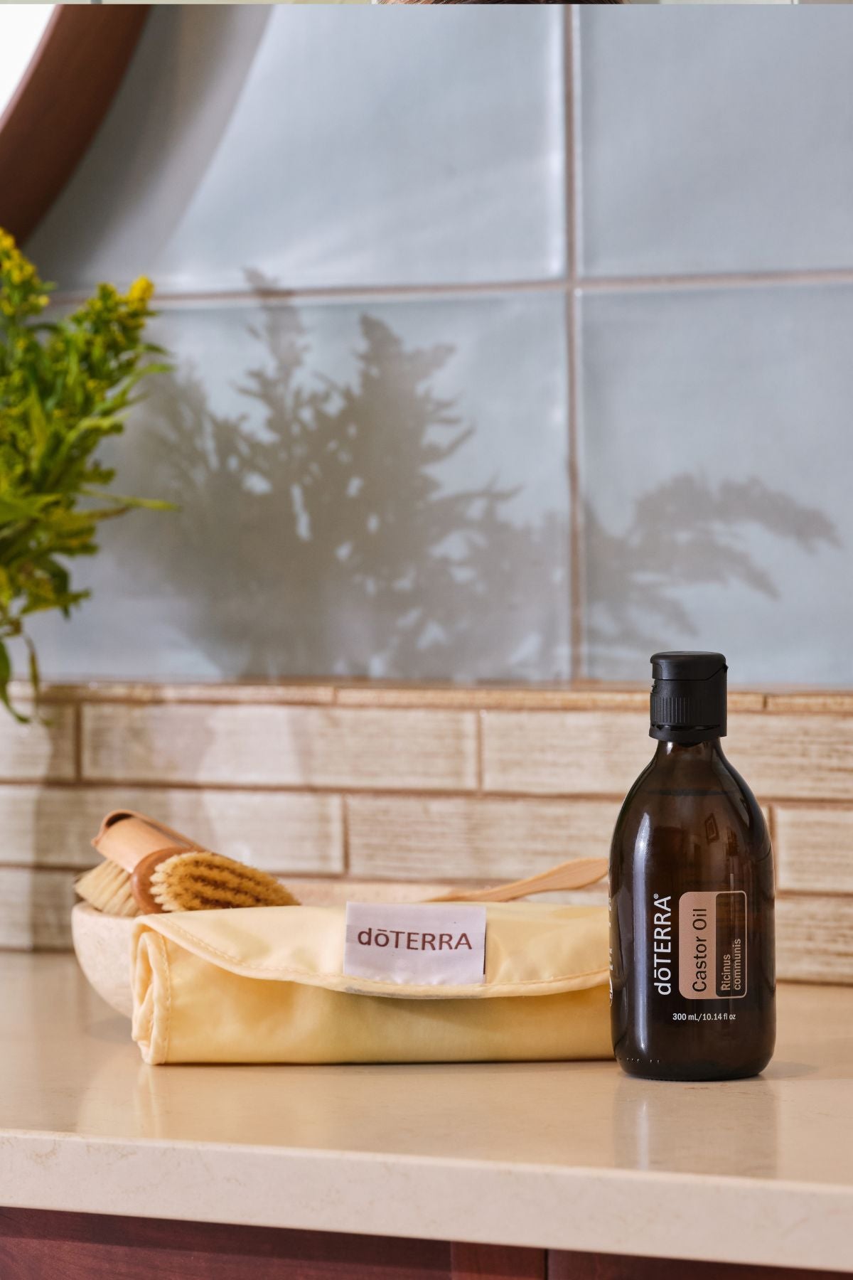 A bottle of doTERRA Castor Oil Wrap Pack oil stands on a countertop next to a folded doTERRA pouch, with a wooden brush in a bowl in the background against a tiled wall.