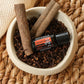 A small black and orange DoTERRA Harvest Spice essential oil bottle rests in a beige bowl filled with cinnamon sticks and cloves, set on a woven straw mat.