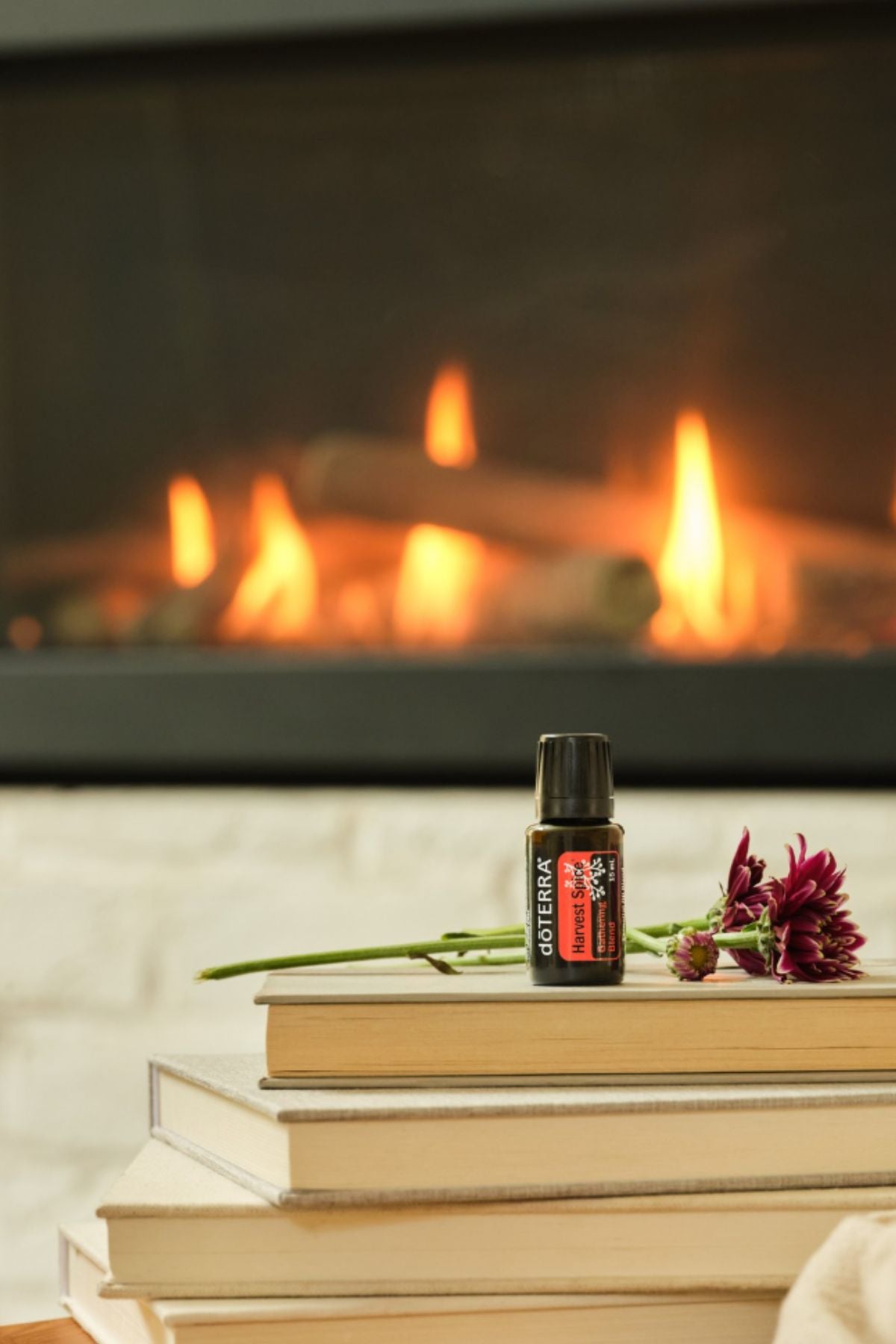 A small bottle of doTERRA Harvest Spice placed on a stack of books in front of a warm fireplace, with a flower resting beside it.