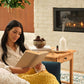 A woman sits in a cozy living room by a fireplace, reading a book wrapped in a chunky mustard yellow knit blanket. A wooden side table beside her holds a white diffuser, a small bowl, and books, with a white brick wall and lit fireplace in the background. doTERRA Harvest Spice