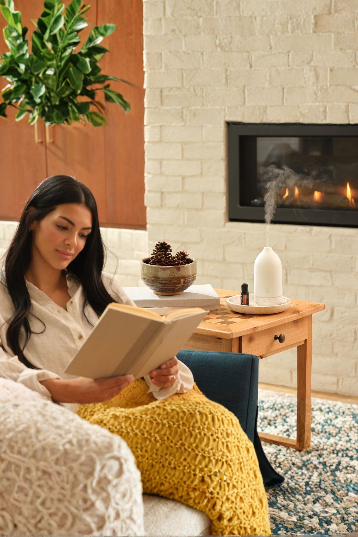 A woman sits in a cozy living room by a fireplace, reading a book wrapped in a chunky mustard yellow knit blanket. A wooden side table beside her holds a white diffuser, a small bowl, and books, with a white brick wall and lit fireplace in the background. doTERRA Harvest Spice