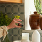 A hand holds a small amber essential oil bottle in a kitchen setting with patterned gray-green tile backsplash, ceramic pots, a white diffuser, and a candle on a woven placemat. doTERRA Harvest Spice