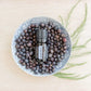 A small black essential oil bottle labeled doTERRA sits on top of a bowl filled with dark dried botanicals, arranged on a light wood surface with green sprigs beside it. doTERRA Juniper Berry Essential Oil
