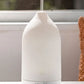 A white essential oil diffuser with misting at the top, sitting on a white surface in front of a window. dōTERRA LaLuz Diffuser