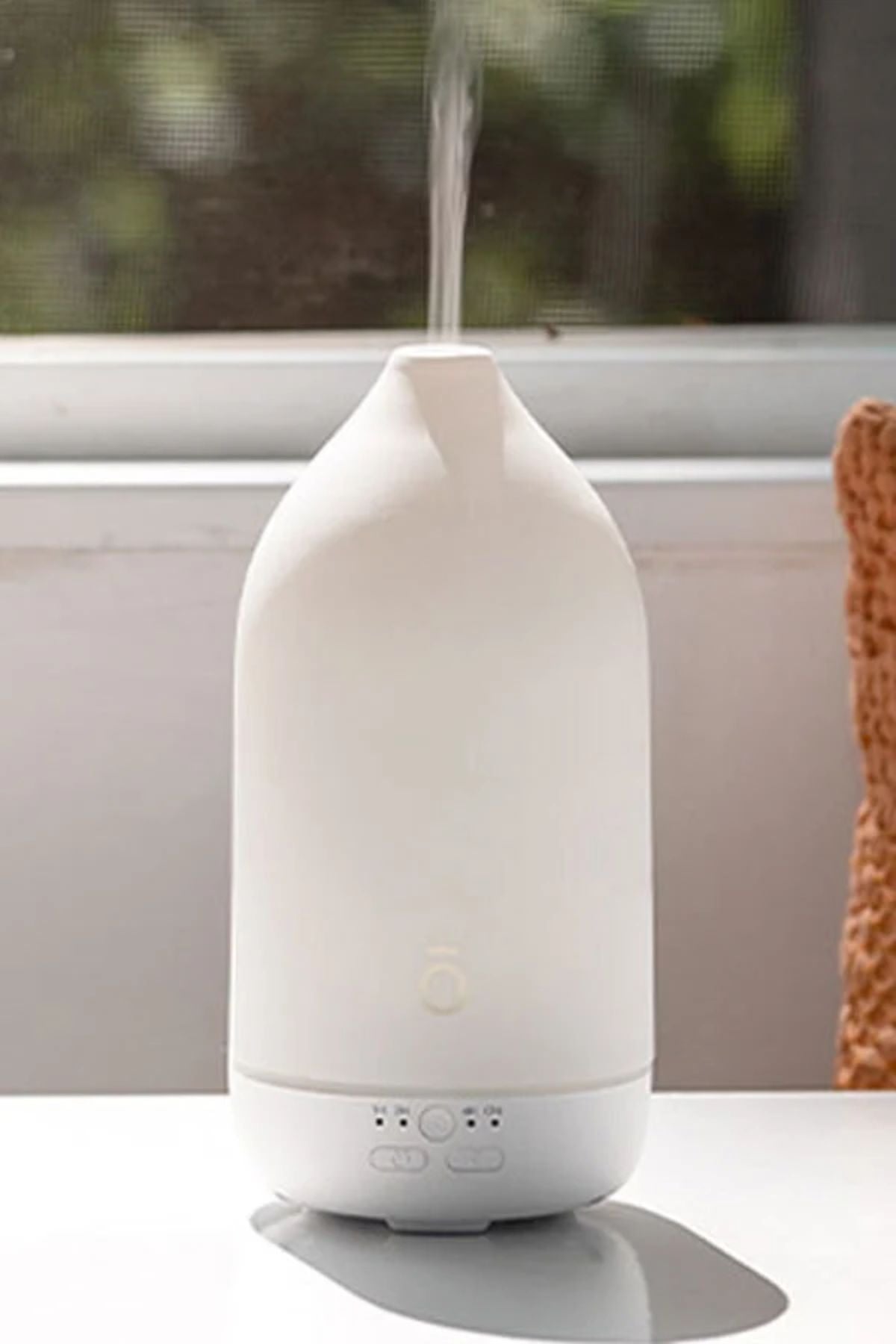 A white essential oil diffuser with misting at the top, sitting on a white surface in front of a window. dōTERRA LaLuz Diffuser