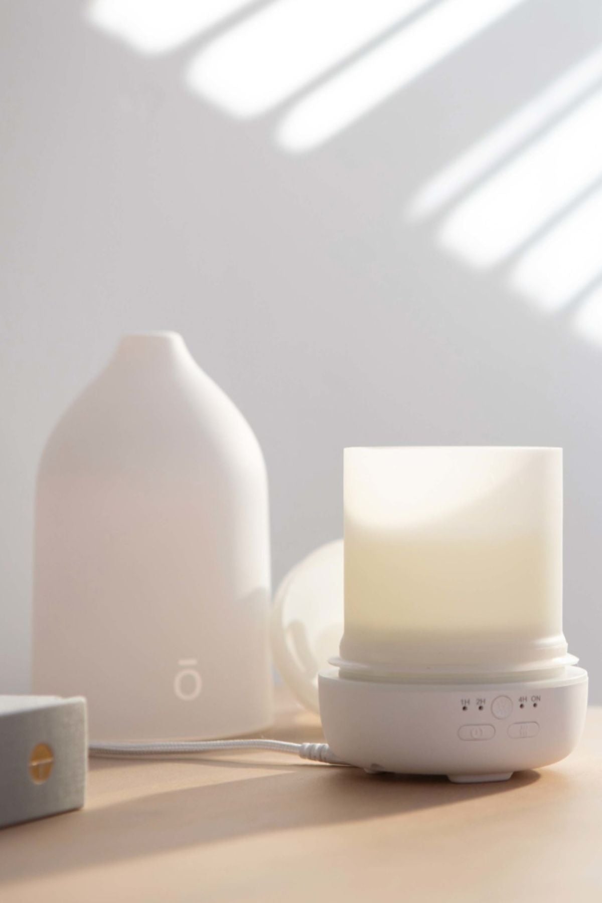A white diffuser and its matching bottle on a light wooden surface, with soft window light casting diagonal shadows on a white wall. dōTERRA LaLuz Diffuser