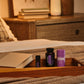 A wooden tray on a bed holds two small dark bottles and a purple tube, with a blanket and a softly blurred bedroom background including a dresser, framed art, and a plant. doTERRA Serenity Sleep System