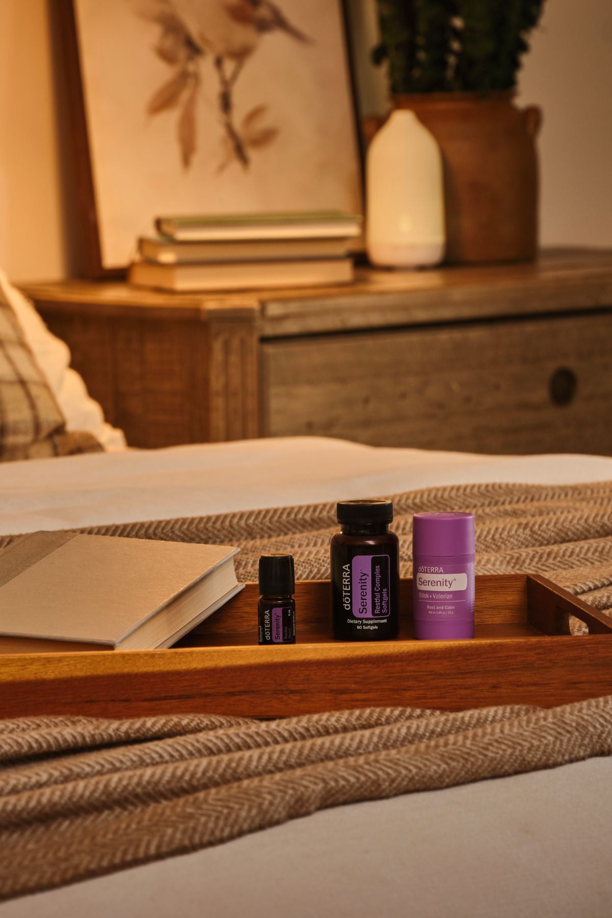 A wooden tray on a bed holds two small dark bottles and a purple tube, with a blanket and a softly blurred bedroom background including a dresser, framed art, and a plant. doTERRA Serenity Sleep System
