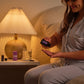 A woman in light gray pajamas sits on a bed at night, holding a small dark bottle over her hand while dispensing a liquid. A bedside table with a lit pleated lamp, a purple bottle, and a glass is visible beside her. doTERRA Serenity Sleep System