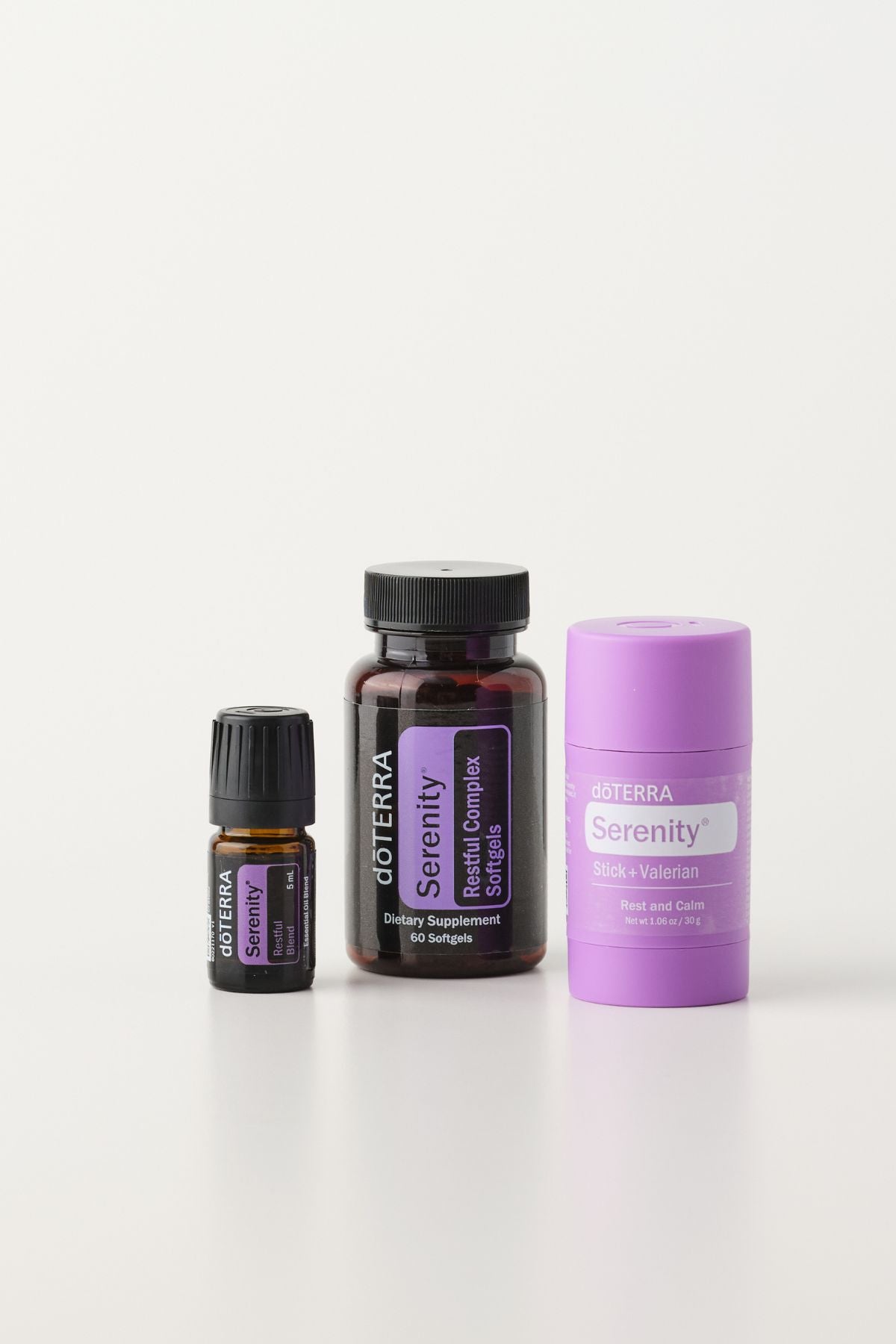 Three doTERRA Serenity products arranged on a clean white studio background: a small dark bottle, a larger amber bottle with a black cap and purple label, and a lavender deodorizer stick. doTERRA Serenity Sleep System