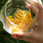 A hand holding a clear glass jar filled with golden-yellow liquid supplement capsules, set against a background of green leaves. Turmeric Dual Chamber Capsules