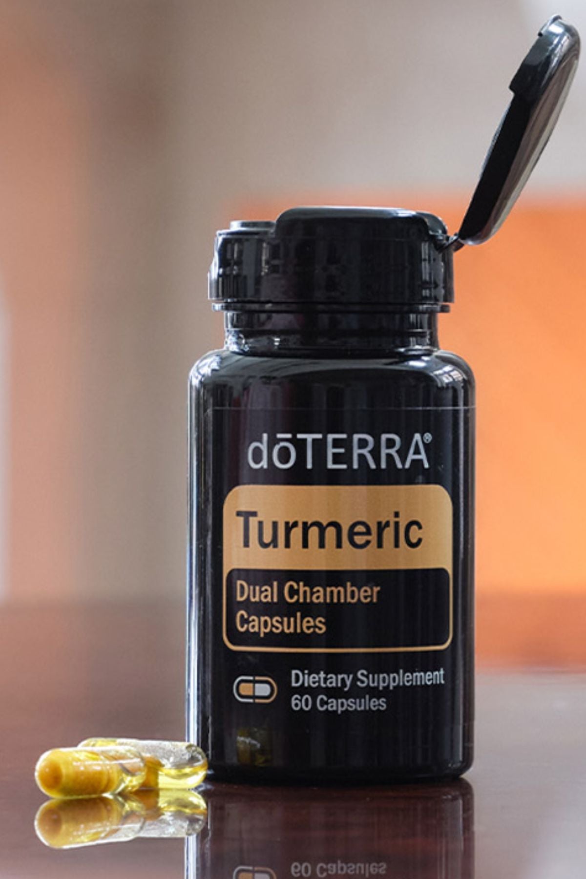 A bottle of doTERRA Turmeric Dual Chamber Capsules with a black cap and label, shown on a reflective surface with a few yellow capsules in front against a warm blurred background.