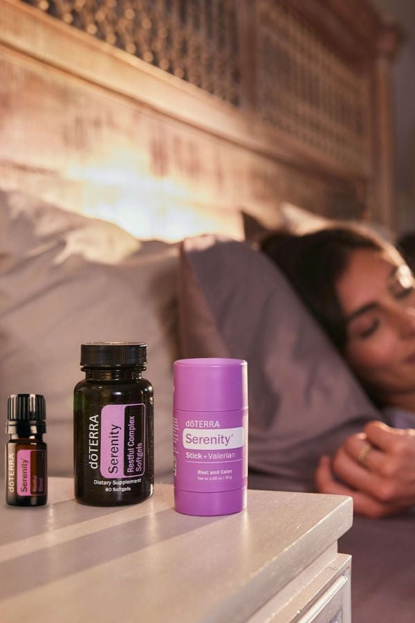A person sleeping in bed in a warm bedroom setting, with three doTERRA Serenity products in the foreground on a bedside table: a small dark bottle, a black bottle, and a purple container. doTERRA Serenity Sleep System